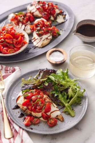 A plate with bruschetta chicken and mixed greens. A drink and platter of more bruschetta chicken and bowl of tomatoes in the background.