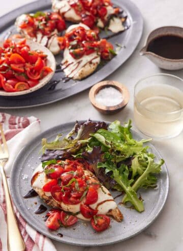 A plate with bruschetta chicken and mixed greens. A drink and platter of more bruschetta chicken and bowl of tomatoes in the background.