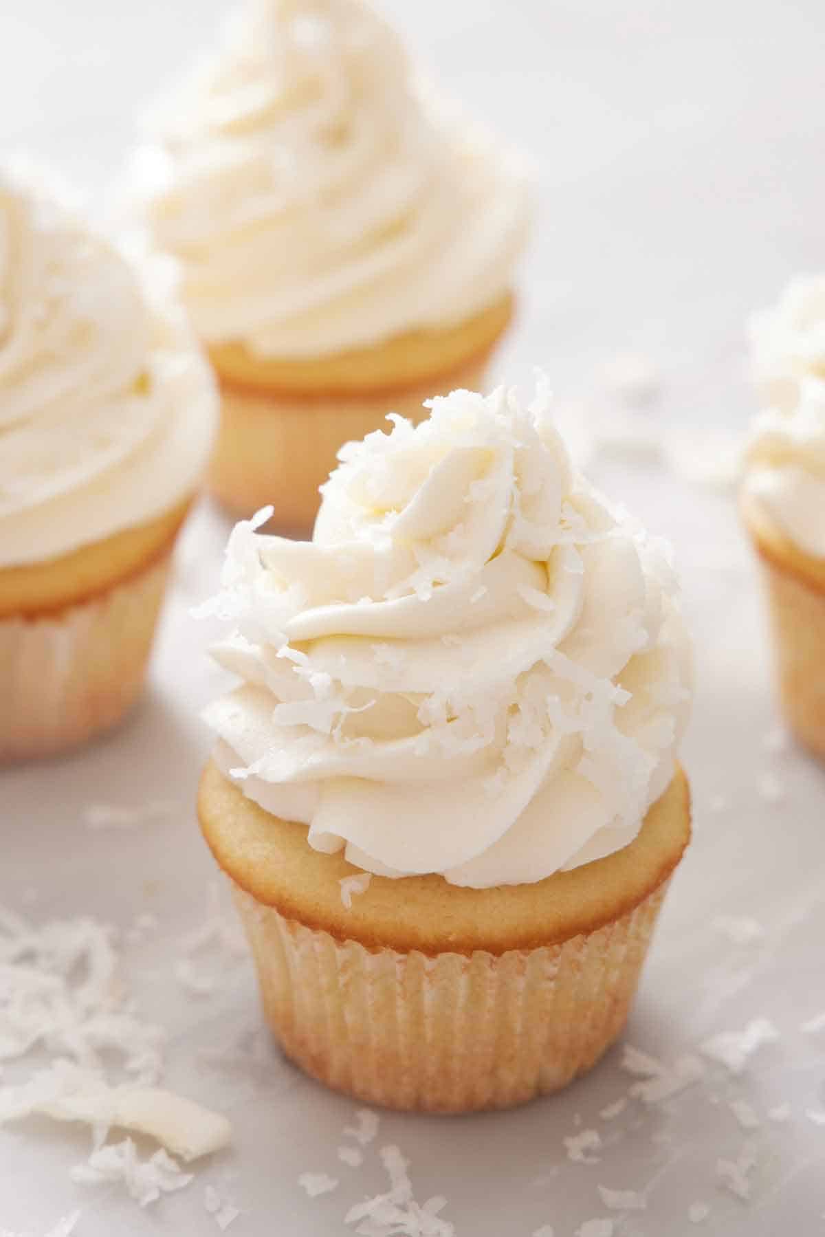 Multiple cupcakes topped with coconut frosting and shaved coconut with one cupcake in focus.