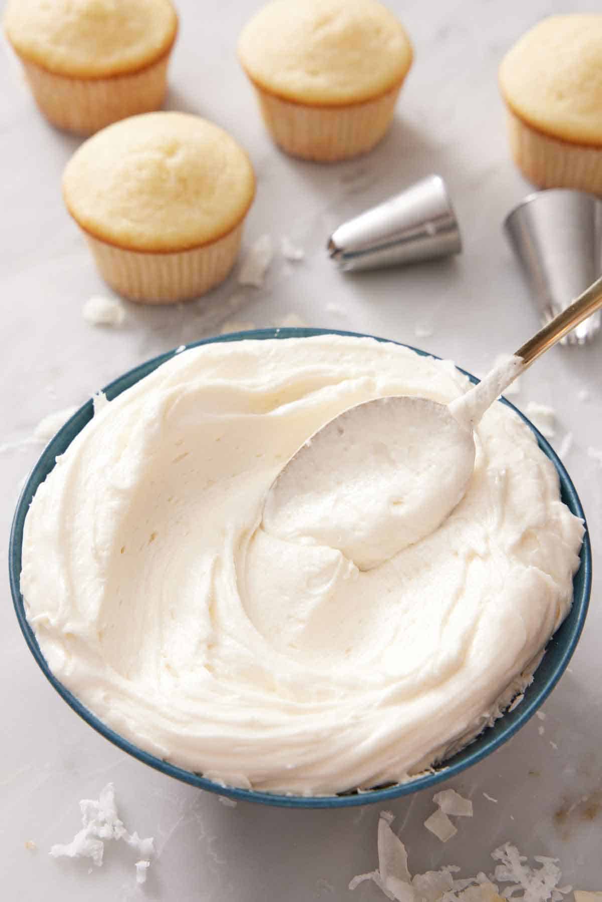 A bowl of coconut frosting with a spoon spread it. Unfrosted cupcakes in the background with piping tips.