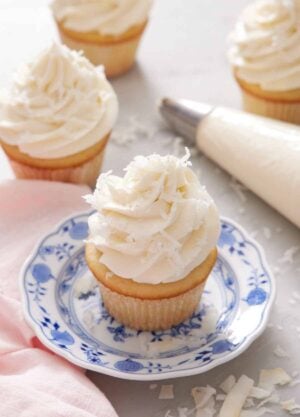 A cupcake topped with coconut frosting on a plate. More frosted cupcakes in the background with a piping bag.