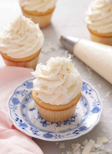 A cupcake topped with coconut frosting on a plate. More frosted cupcakes in the background with a piping bag.