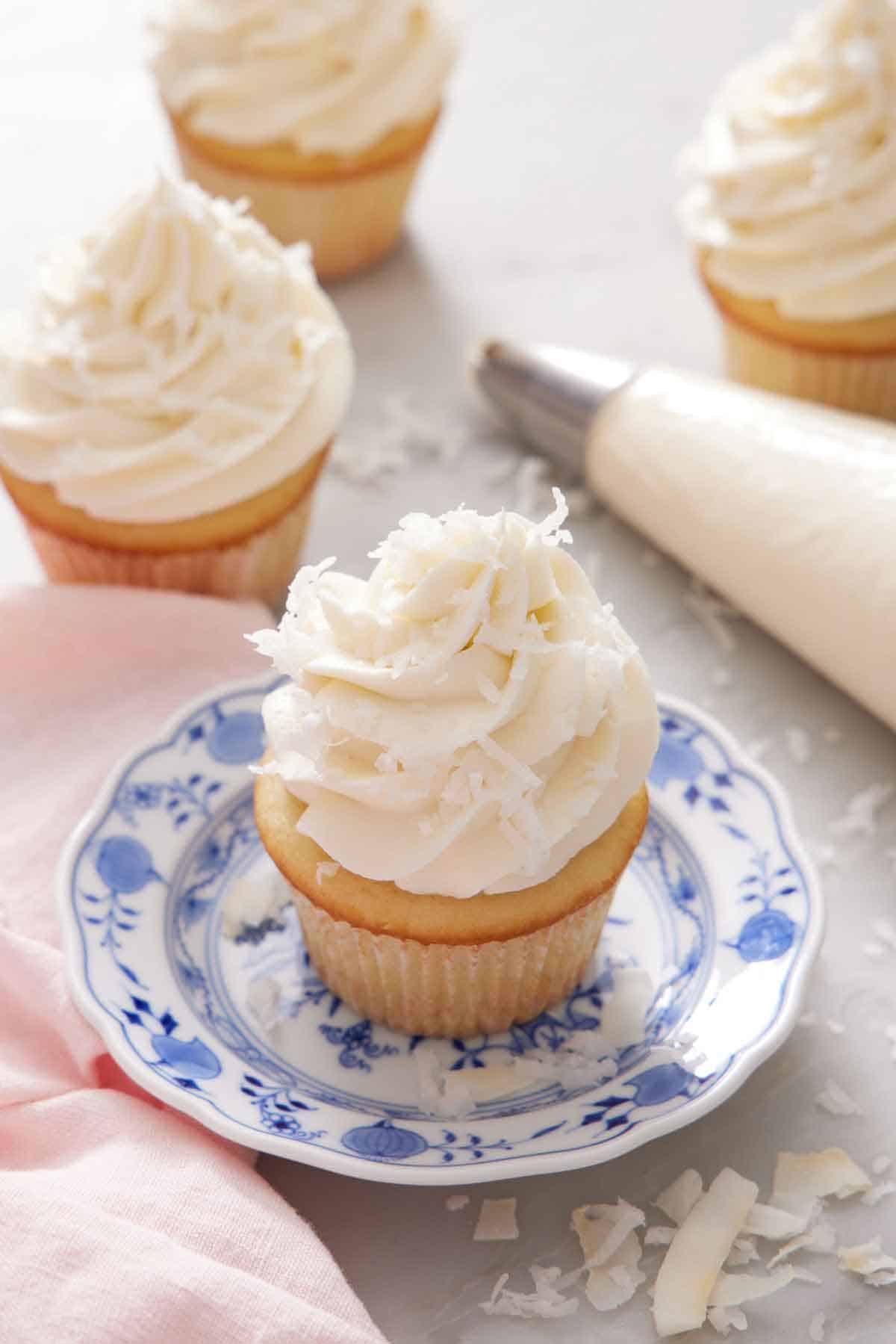 A cupcake topped with coconut frosting on a plate. More frosted cupcakes in the background with a piping bag.