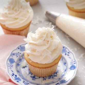 Pinterest graphic of a cupcake topped with coconut frosting on a plate. A a piping bag and more frosted cupcakes in the background.