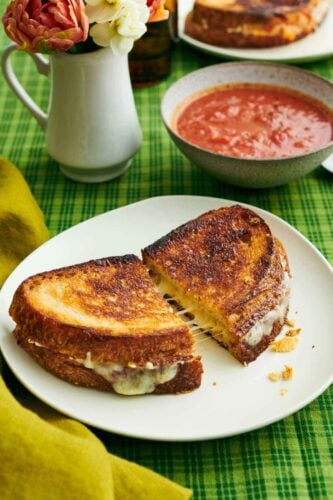 A plate with a grilled cheese cut in half and slightly pulled apart. A bowl of tomato soup and vase of flowers in the back.