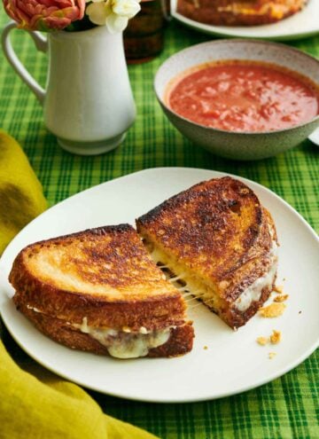 A plate with a grilled cheese cut in half and slightly pulled apart. A bowl of tomato soup and vase of flowers in the back.