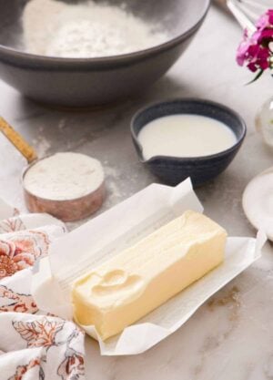 A stick of softened butter in an opened wrapper. A bowl and measuring cup of flour and dairy in the background.