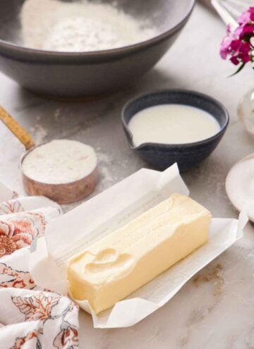 A stick of softened butter in an opened wrapper. A bowl and measuring cup of flour and dairy in the background.