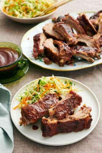 A plate with three pieces of Instant Pot ribs with slaw. A platter of more ribs in the back also with a bowl of sauce.