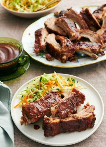 A plate with three pieces of Instant Pot ribs with slaw. A platter of more ribs in the back also with a bowl of sauce.