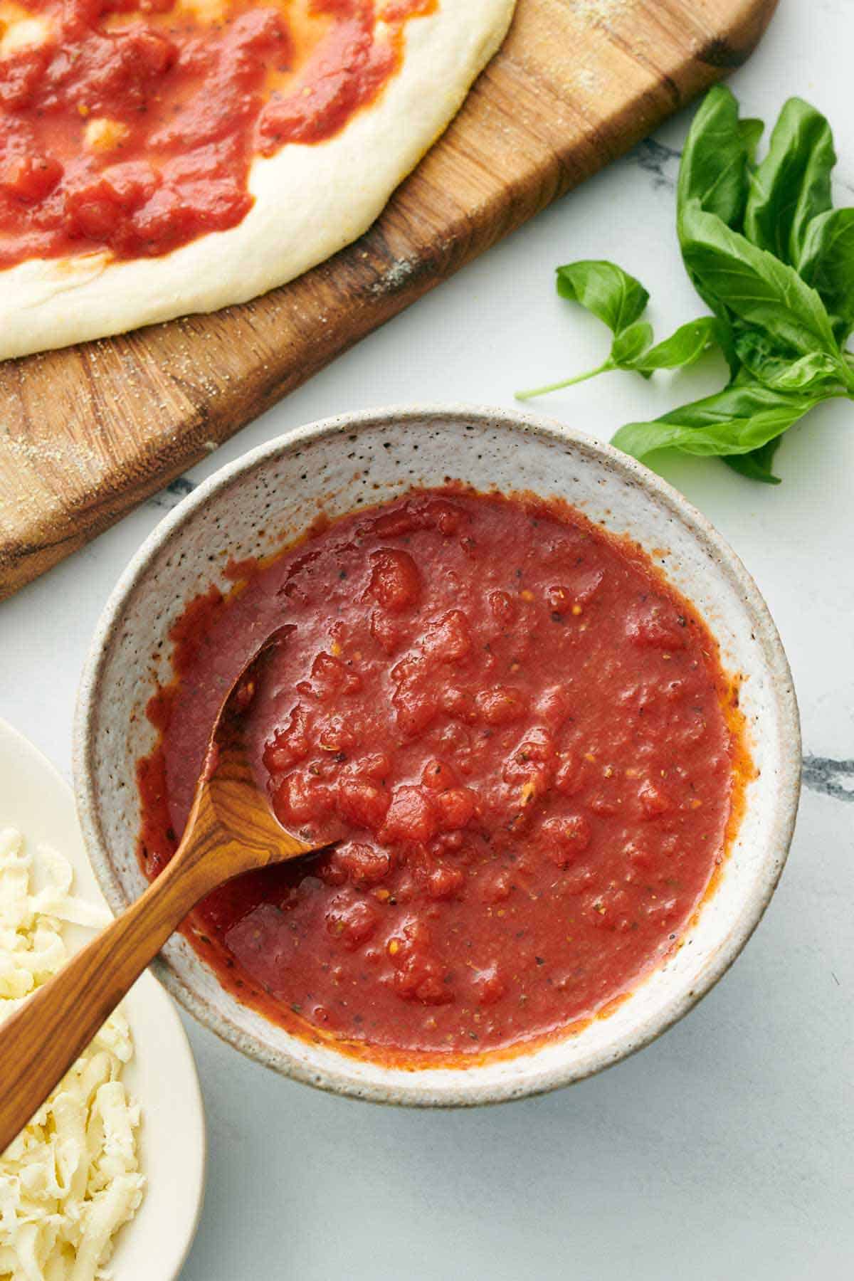 Overhead view of a bowl of pizza sauce with a wooden spoon. Fresh basil and pizza dough with sauce spread on in the background.