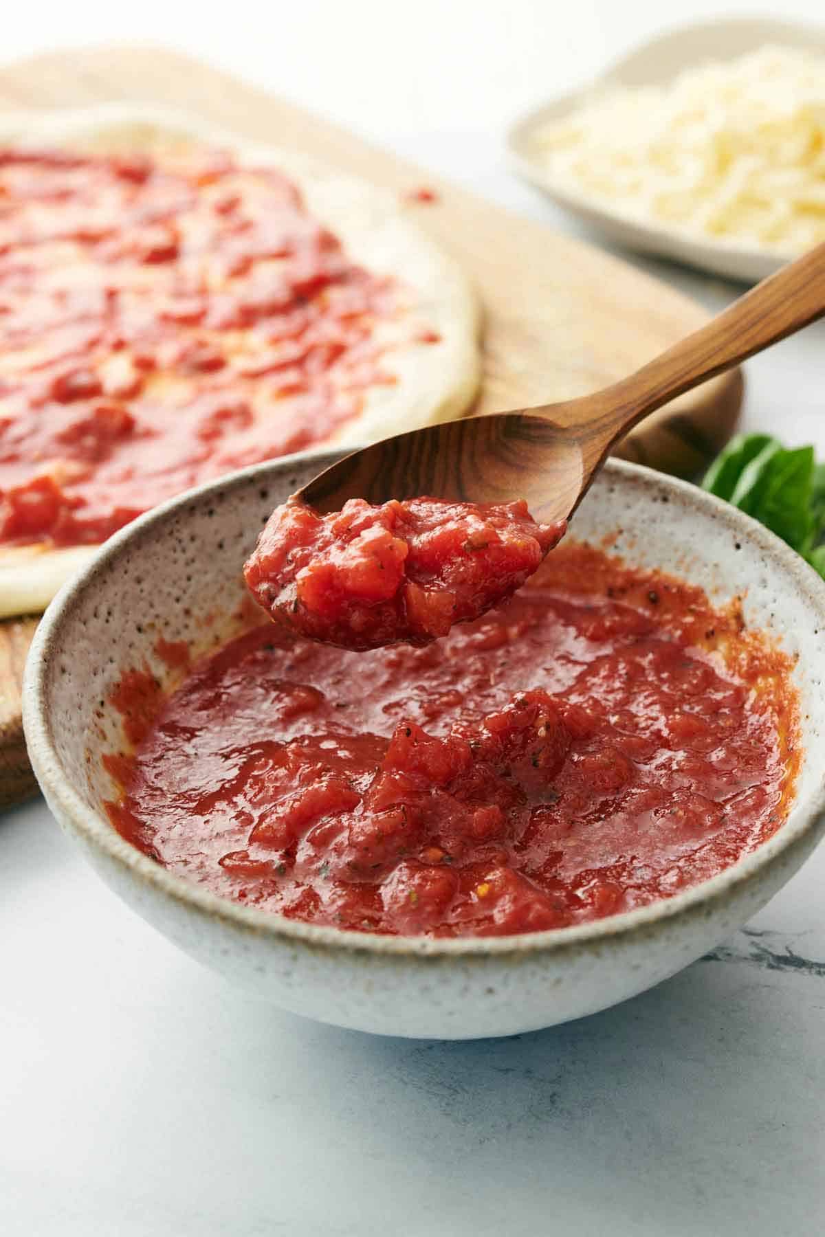 A spoon lifting up homemade pizza sauce from a bowl. Pizza dough with sauce on top in the background.