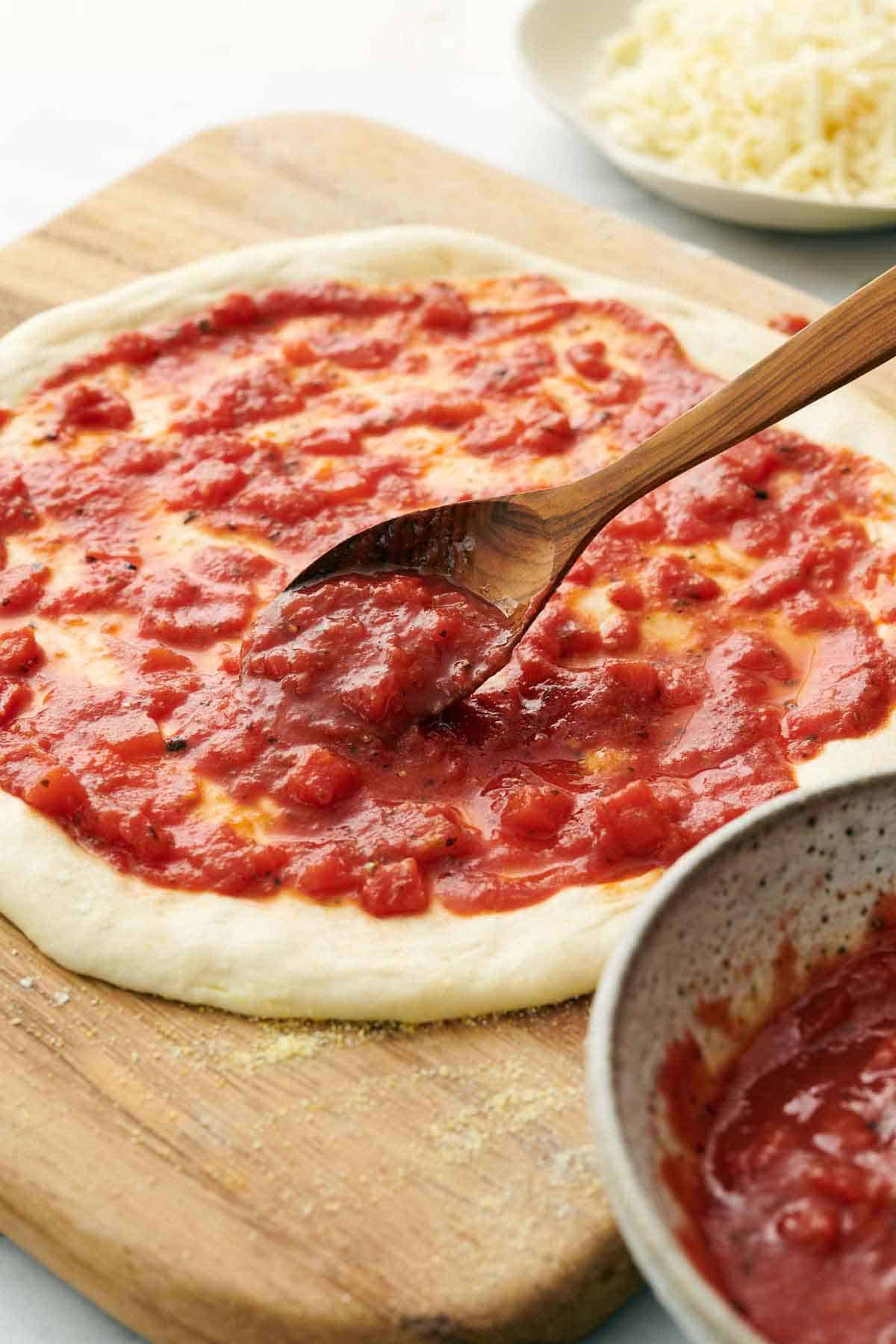 A spoon adding pizza sauce to stretched pizza dough.