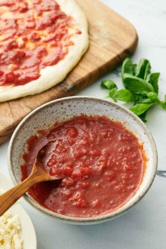 A bowl of homemade pizza sauce with a spoon. Pizza dough with sauce spread on top in the background along with fresh basil on the counter.