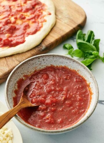 A bowl of homemade pizza sauce with a spoon. Pizza dough with sauce spread on top in the background along with fresh basil on the counter.