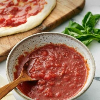 Pinterest graphic of a bowl of homemade pizza sauce with a spoon. Pizza dough with sauce spread on top in the background along with fresh basil.