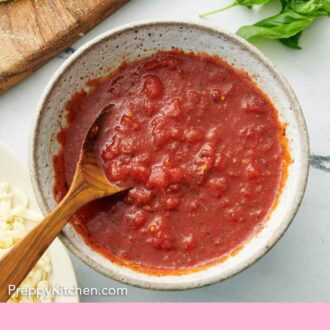 Pinterest graphic of overhead view of a bowl of pizza sauce with a wooden spoon. Fresh basil in the background.