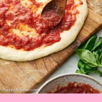 Pinterest graphic of a spoon adding homemade pizza sauce to stretched pizza dough on a wooden board. Fresh basil on the side.