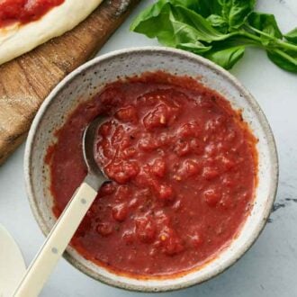 Overhead view of a bowl of pizza sauce with a spoon inside. Pizza ingredients surrounding it.