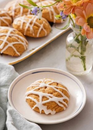 A plate with a pumpkin cookie with icing drizzled on top. A platter of more pumpkin cookies and a vase of flower in the back.
