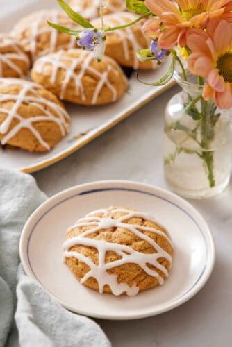 A plate with a pumpkin cookie with icing drizzled on top. A platter of more pumpkin cookies and a vase of flower in the back.