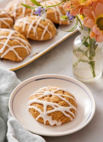 A plate with a pumpkin cookie with icing drizzled on top. A platter of more pumpkin cookies and a vase of flower in the back.