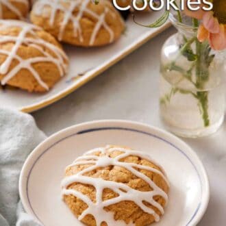 Pinterest graphic of a plate with a pumpkin cookie with icing drizzled on top. A platter of more pumpkin cookies and a vase of flower in the back.