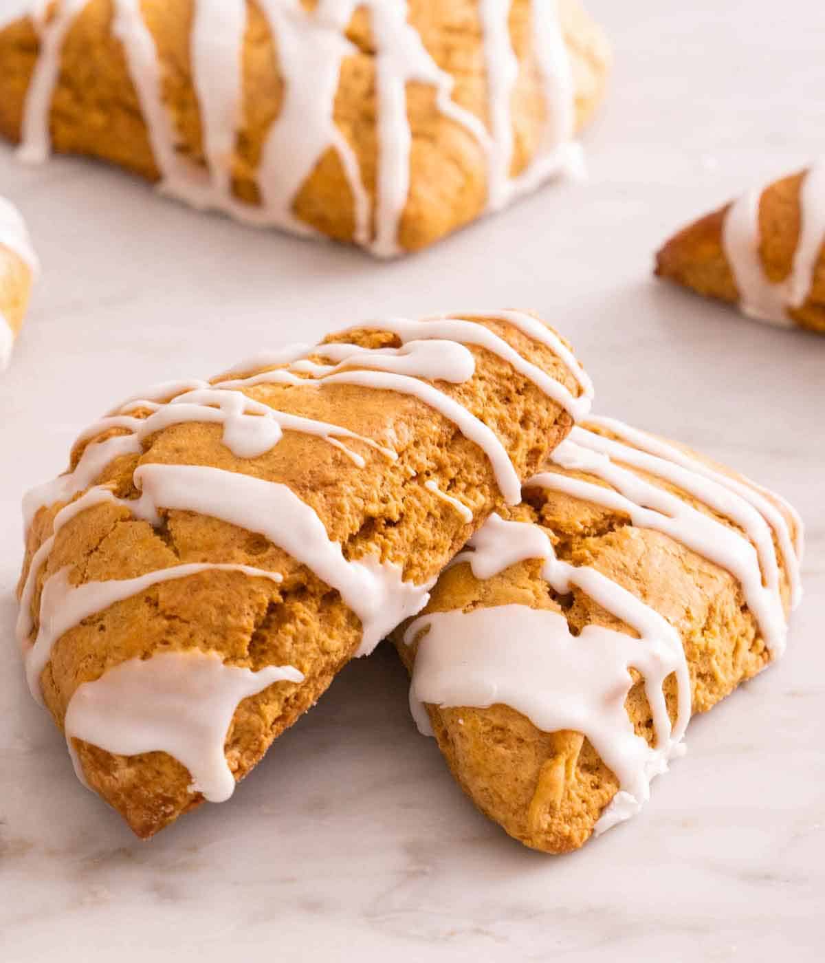 Two pumpkin scones with icing drizzled on top, with one slightly stacked on top. More scones in the background.