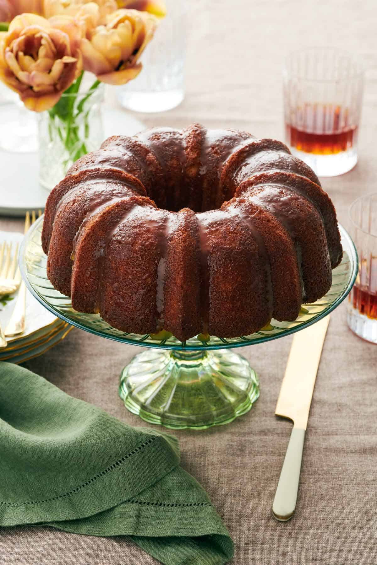 A green cake stand with a rum cake on top. A knife, linen napkin, drinks, and flowers beside it.