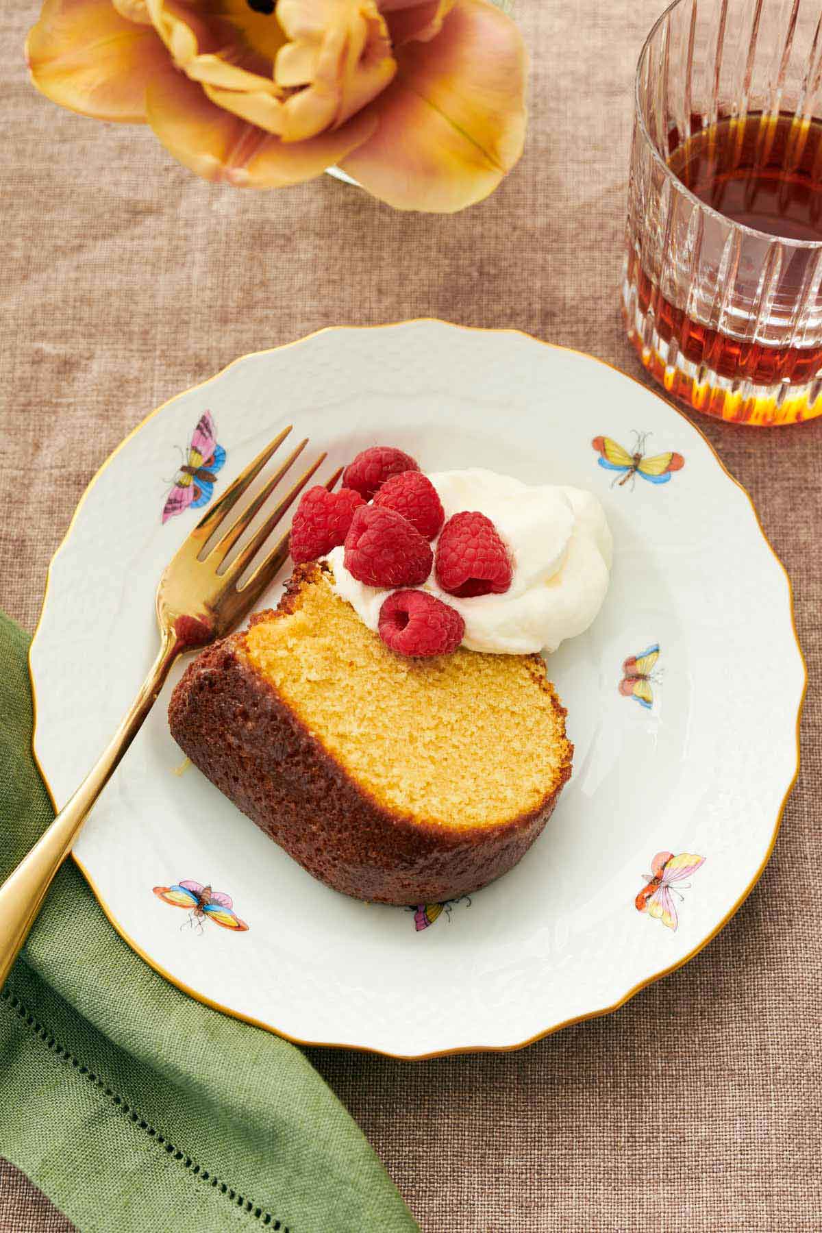 A plate with a slice of rum cake with whipped cream, raspberries, and a fork. A drink beside it.