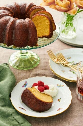 A slice of rum cake on a plate with whipped cream and raspberries. The rest of the cake on a cake stand behind it along with stacks of plates and forks.