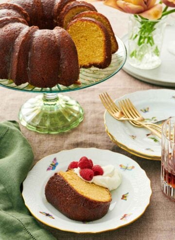 A slice of rum cake on a plate with whipped cream and raspberries. The rest of the cake on a cake stand behind it along with stacks of plates and forks.