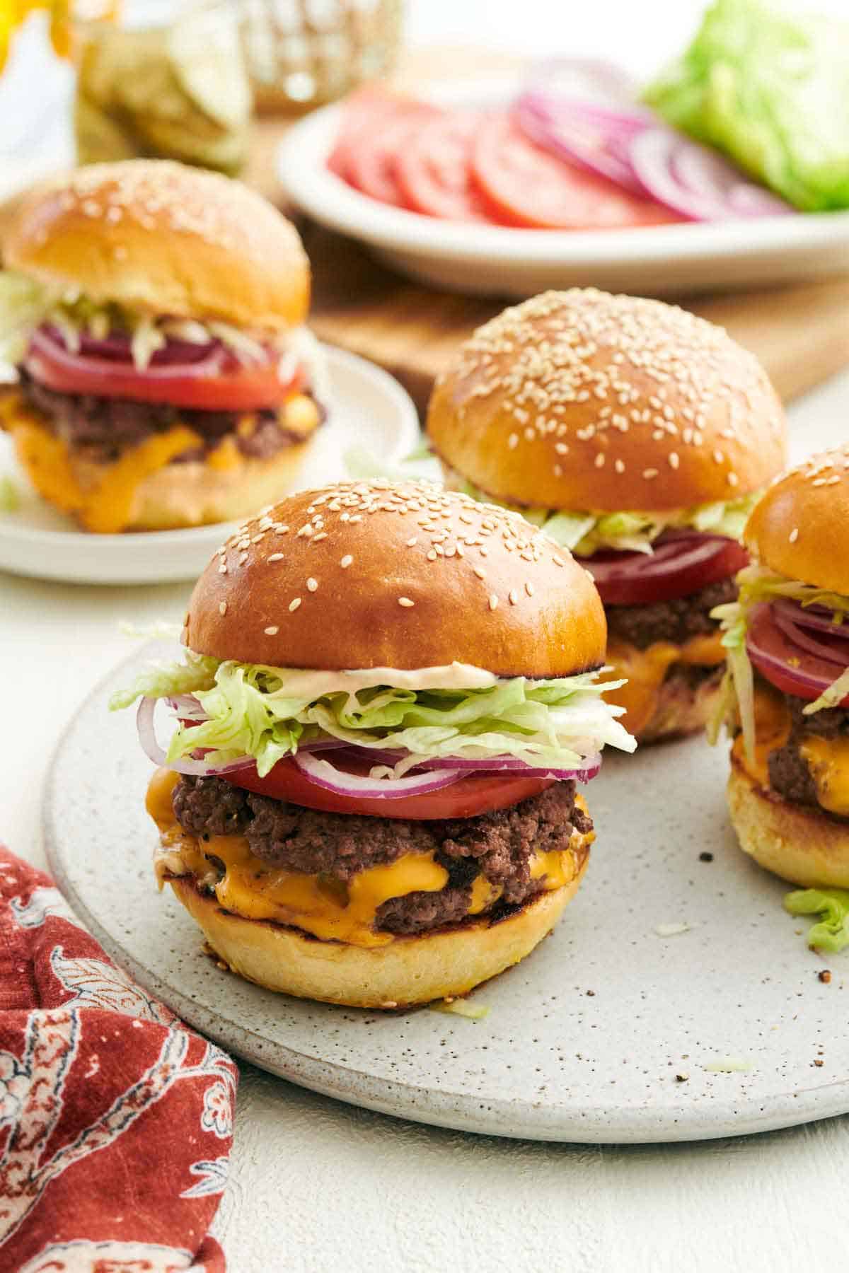 Multiple smash burgers on a platter and one on a plate in the background. A platter with burger toppings in the back.