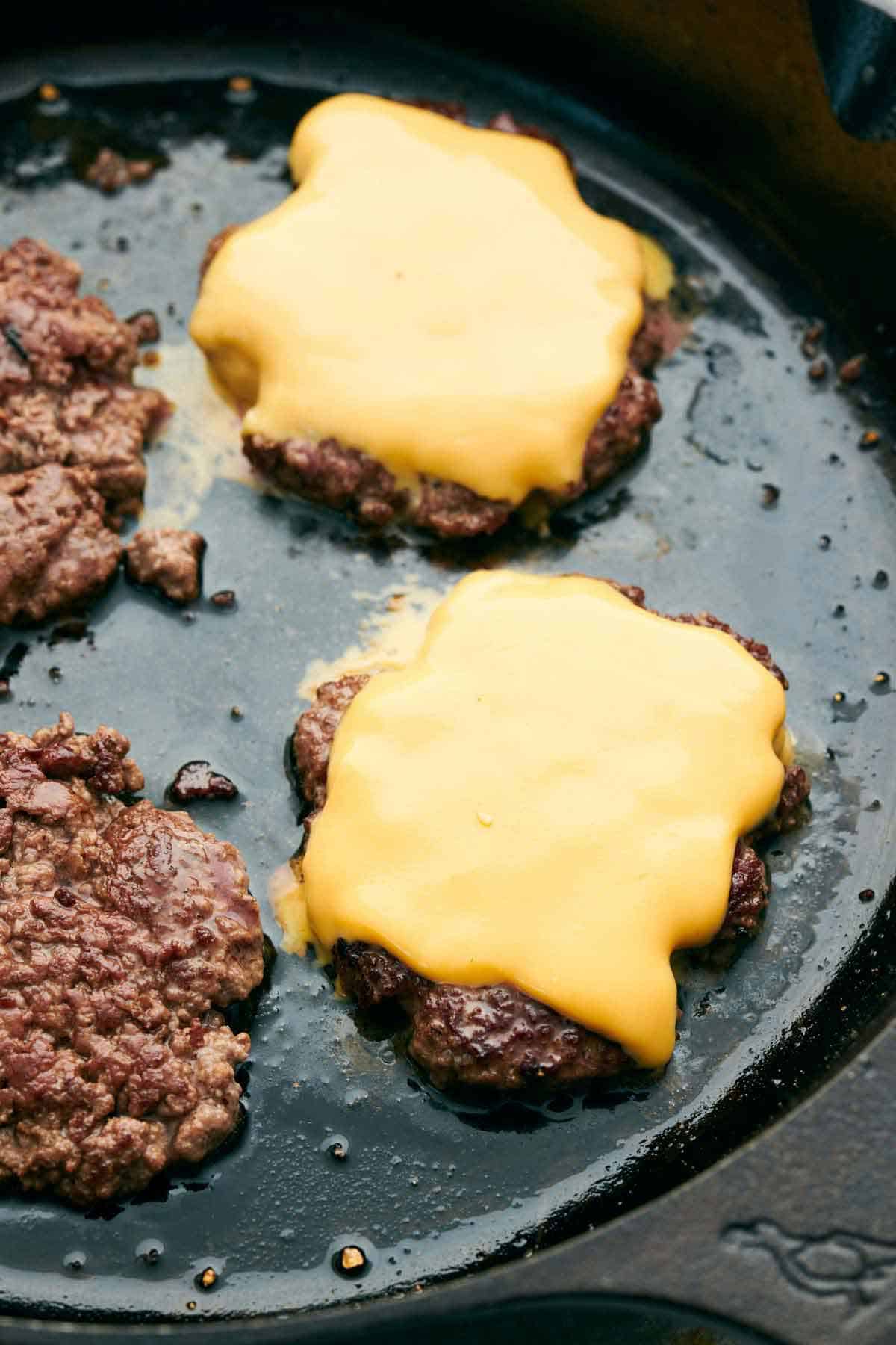 Smash burger patties on a cast iron grill, two patties with sliced cheddar cheese on top.