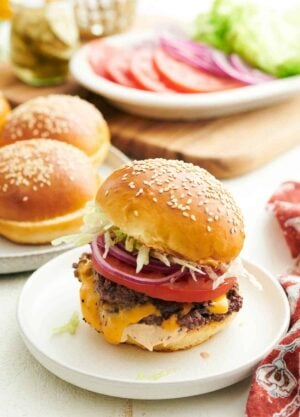 A smash burger on a plate with buns in the background along with tomatoes, onions, and lettuce.