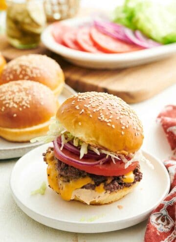 A smash burger on a plate with buns in the background along with tomatoes, onions, and lettuce.