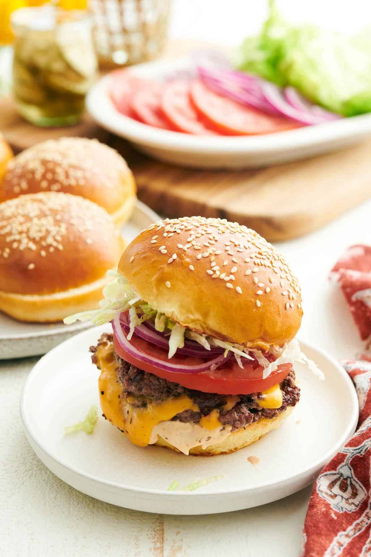 A smash burger on a plate with buns in the background along with tomatoes, onions, and lettuce.