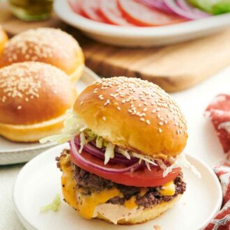 Pinterest graphic of a smash burger on a plate with buns in the background along with tomatoes, onions, and lettuce.