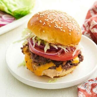 A smash burger topped with a slice of tomato, onions, and lettuce on a plate. A napkin on the side along with a platter of toppings.