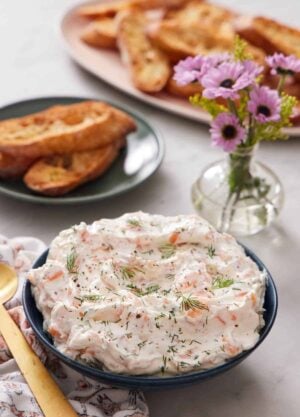 A bowl of smoked salmon dip topped with fresh dill. Bread and a vase of flowers in the background.