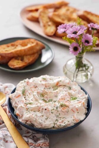 A bowl of smoked salmon dip topped with fresh dill. Bread and a vase of flowers in the background.