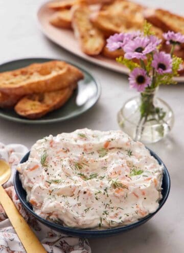 A bowl of smoked salmon dip topped with fresh dill. Bread and a vase of flowers in the background.