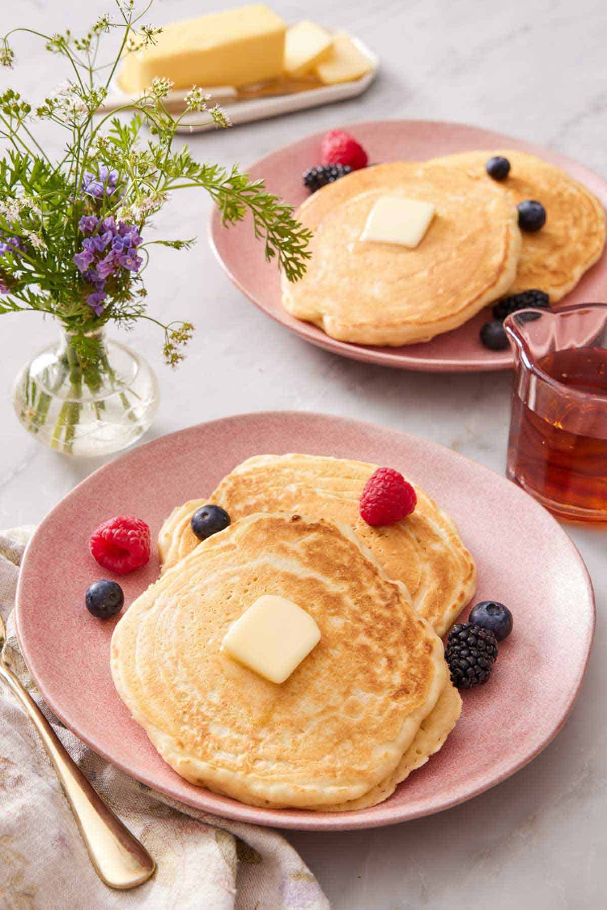 A plate with two sourdough pancakes topped with berries and butter. A second plate in the background along with syrup.