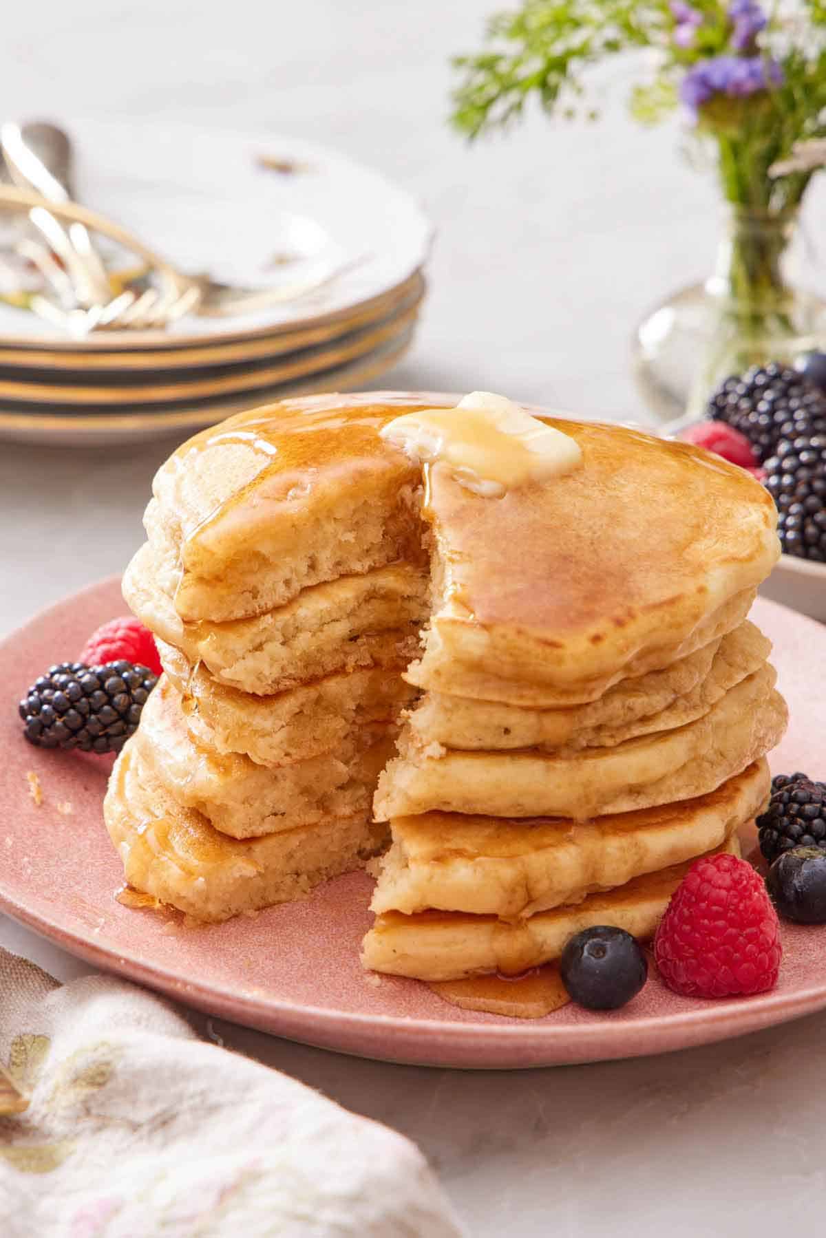 A stack of cut sourdough pancakes on a plate with syrup running down the sides and butter on top. Berries on the plate and a stack of plates in the background.