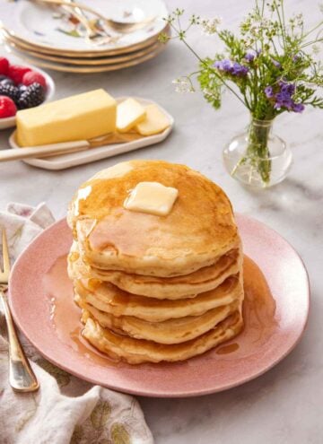 A pink plate with a stack of sourdough pancakes topped with butter and maple syrup. A platter of butter, vase of flowers, and stack of plates in the background.