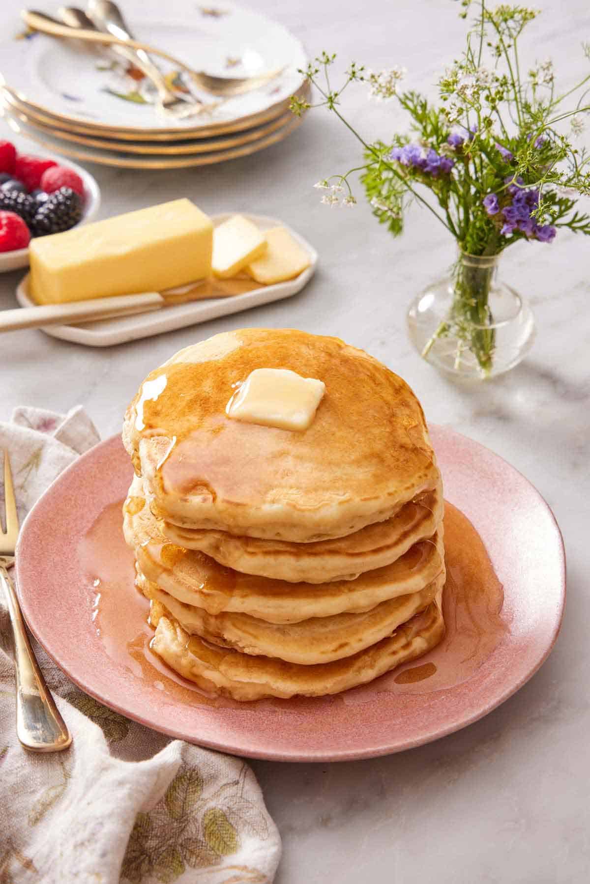 A pink plate with a stack of sourdough pancakes topped with butter and maple syrup. A platter of butter, vase of flowers, and stack of plates in the background.