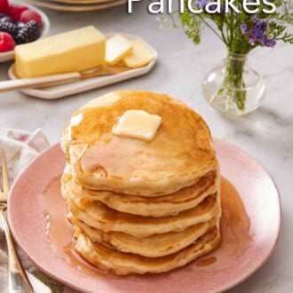 Pinterest graphic of a plate with a stack of sourdough pancakes topped with butter and maple syrup. Butter, vase of flowers, and stack of plates in the background.