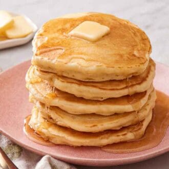 A stack of sourdough pancakes with butter and syrup on top. Butter in the background and a fork beside the plate.