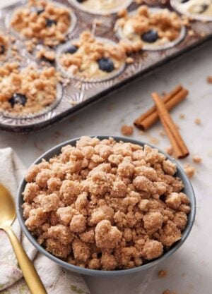 A bowl of streusel topping with cinnamon sticks and muffins topped with streusel in the background.