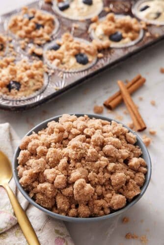 A bowl of streusel topping with cinnamon sticks and muffins topped with streusel in the background.